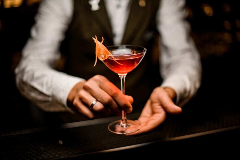 Close-up view of wine glass with trendy bright alcoholic cocktail in hands of male bartender. Blurred background